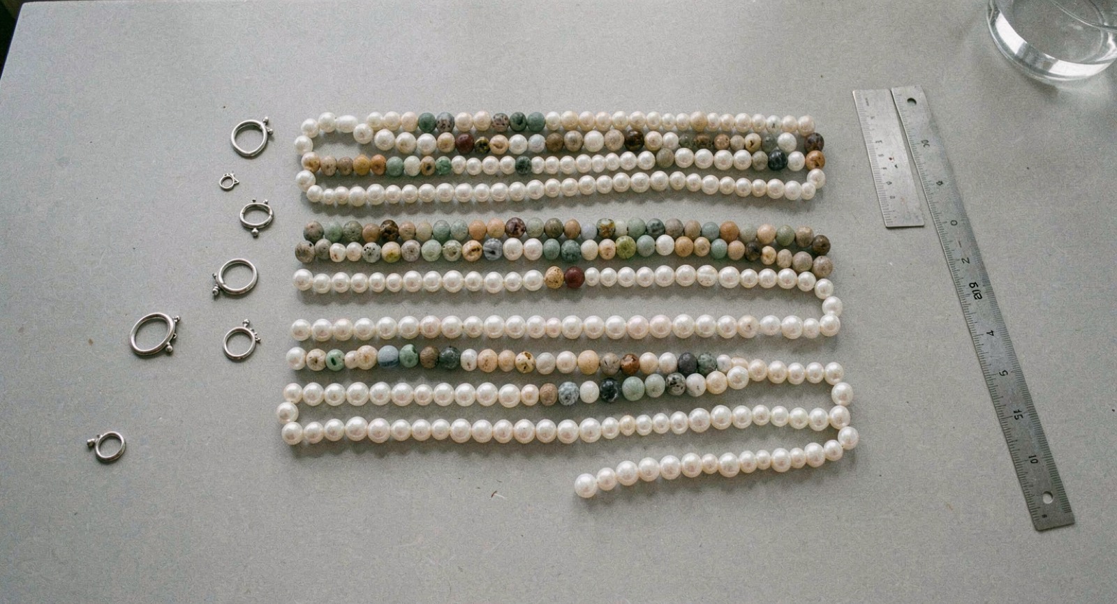 Flere strenge hvide perler og brogede stenperler lagt i parallelle rækker på et lysegråt bord, med små sølvlåse og en stållineal ved siden af, set lige ovenfra i dagslys